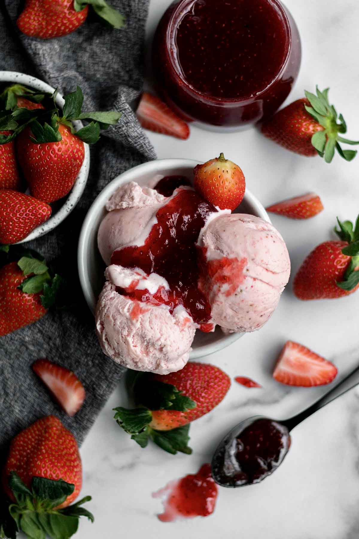 A bowl of Strawberry Ice Cream sits with chopped strawberries strewn on the table with a ramekin of Strawberry Sauce eyeing the scene.