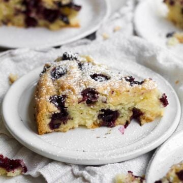 A slice of gluten free olive oil cake dusted with confectioners’ sugar dotted by bursted blueberries.
