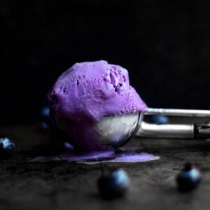 A purple ball of gluten free Blueberry Ice Cream sits in a scoop slightly melting with blueberries on the table.