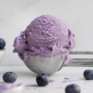 A sphere of vibrant lavender blueberry ice cream rests in a silver scoop above scattered fresh blueberries.