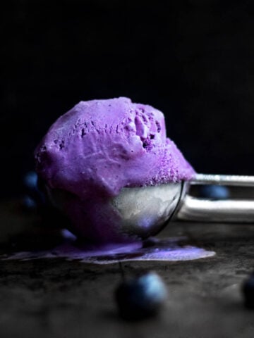 A purple ball of gluten free Blueberry Ice Cream sits in a scoop slightly melting with blueberries on the table.