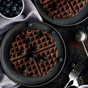 Two circular gluten free Chocolate Waffles on individual black plates topped with blueberries.
