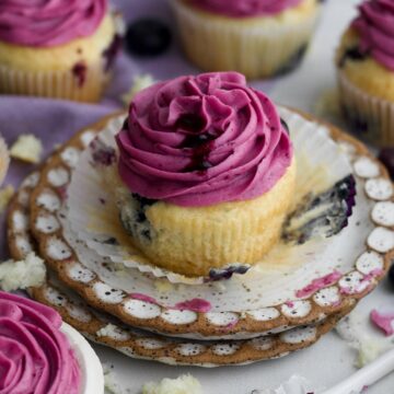 An unwrapped gluten-free blueberry cupcake sits on a plate with creamy magenta blueberry frosting.