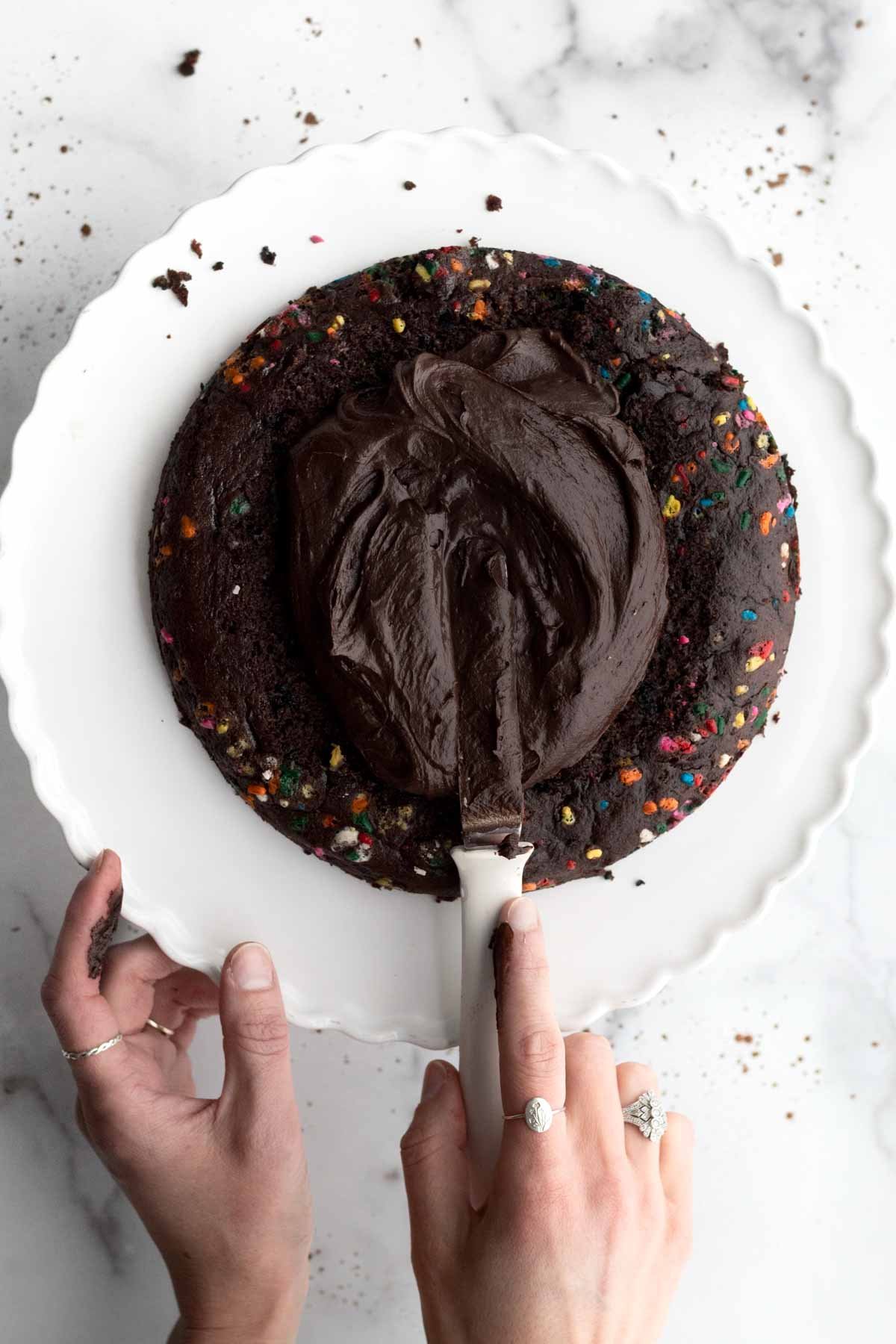 Spreading chocolate fudge frosting on a dark rainbow sprinkled cake.