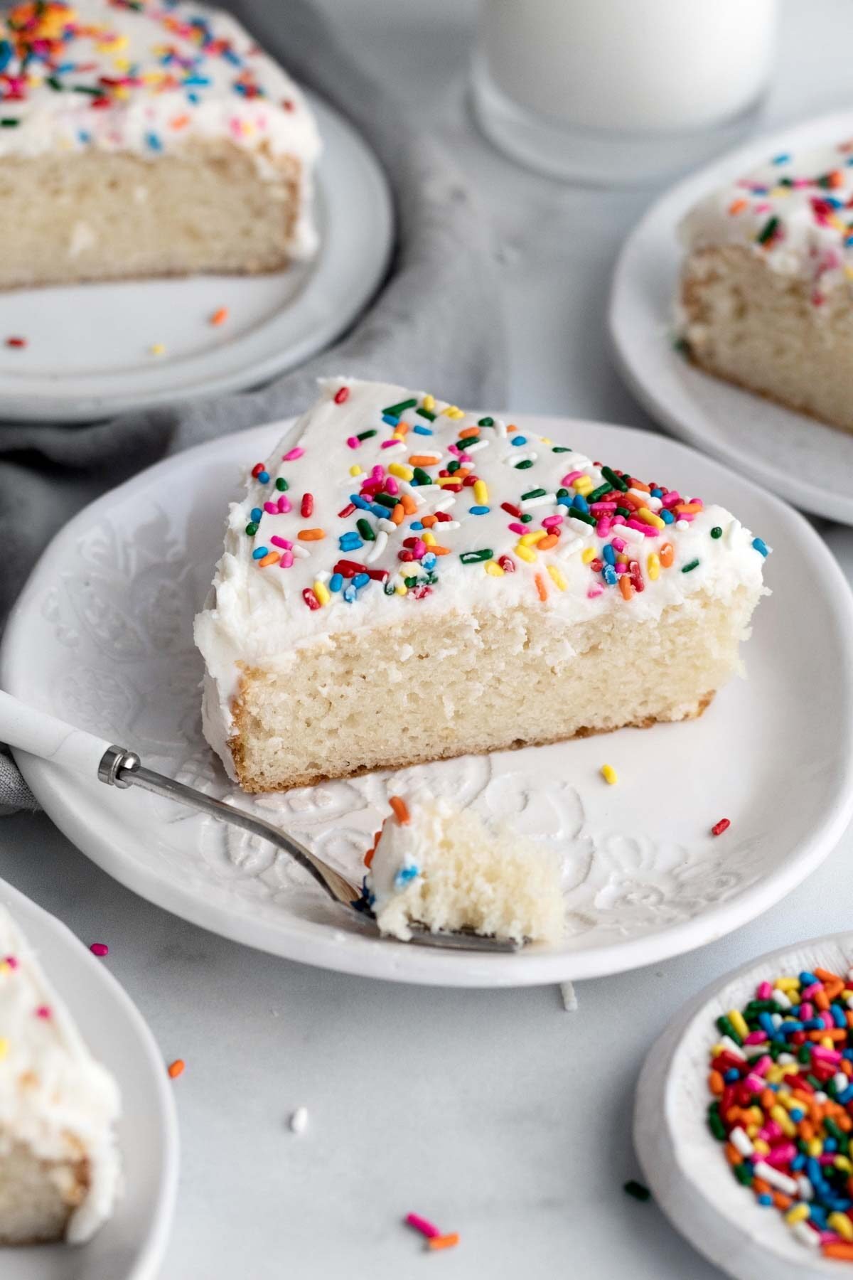A slice of eggless vanilla cake topped with frosting and rainbow sprinkles.