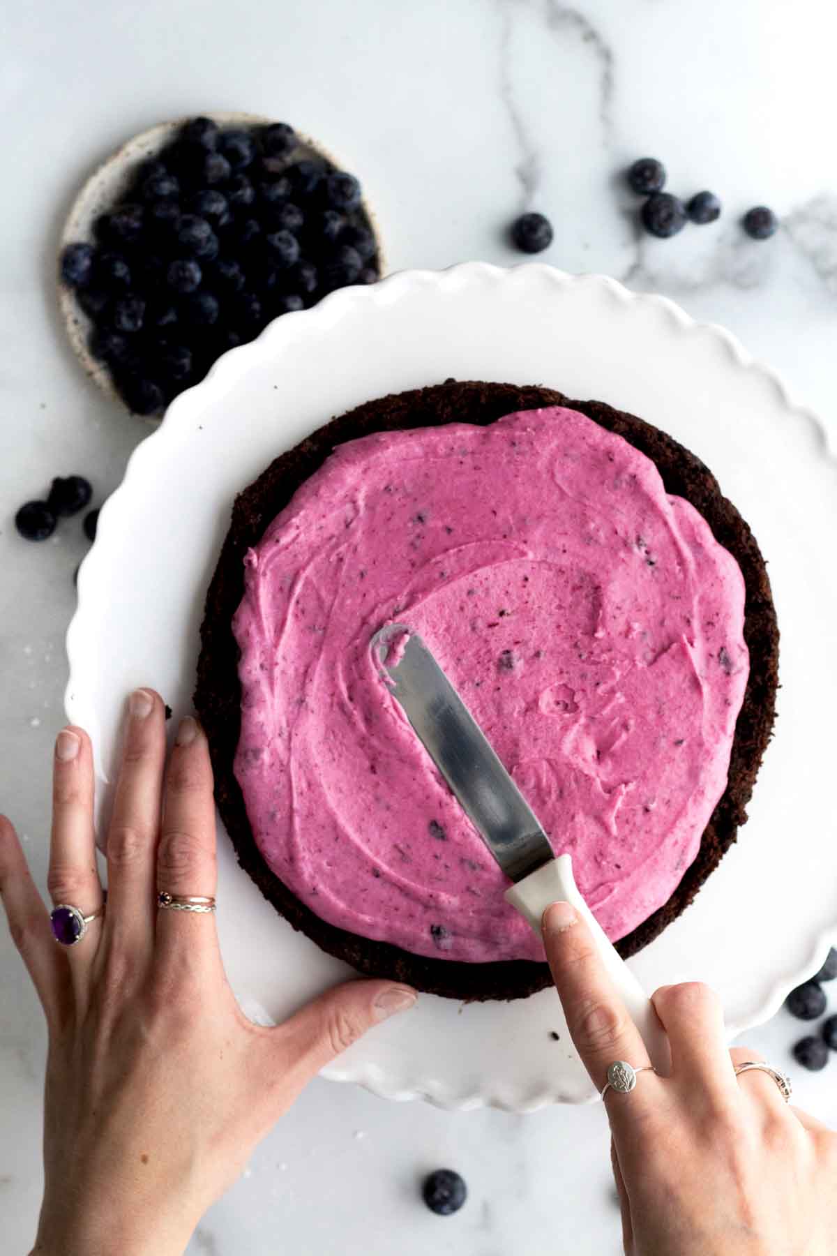 Hands spreading blueberry frosting on a chocolate cake with a spatula.