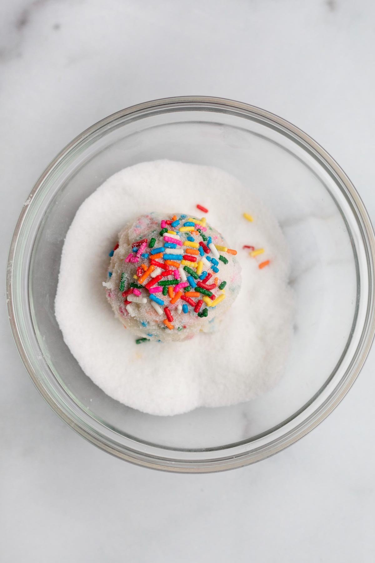 A rainbow sprinkled ball of dough in a bowl of sugar.