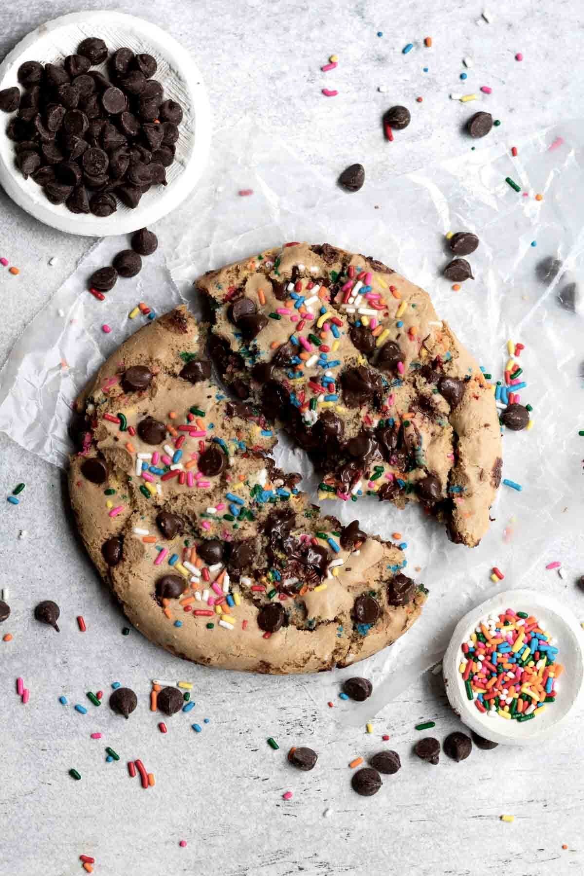 A cookie broken in half with bowls of chocolate chips and sprinkles.