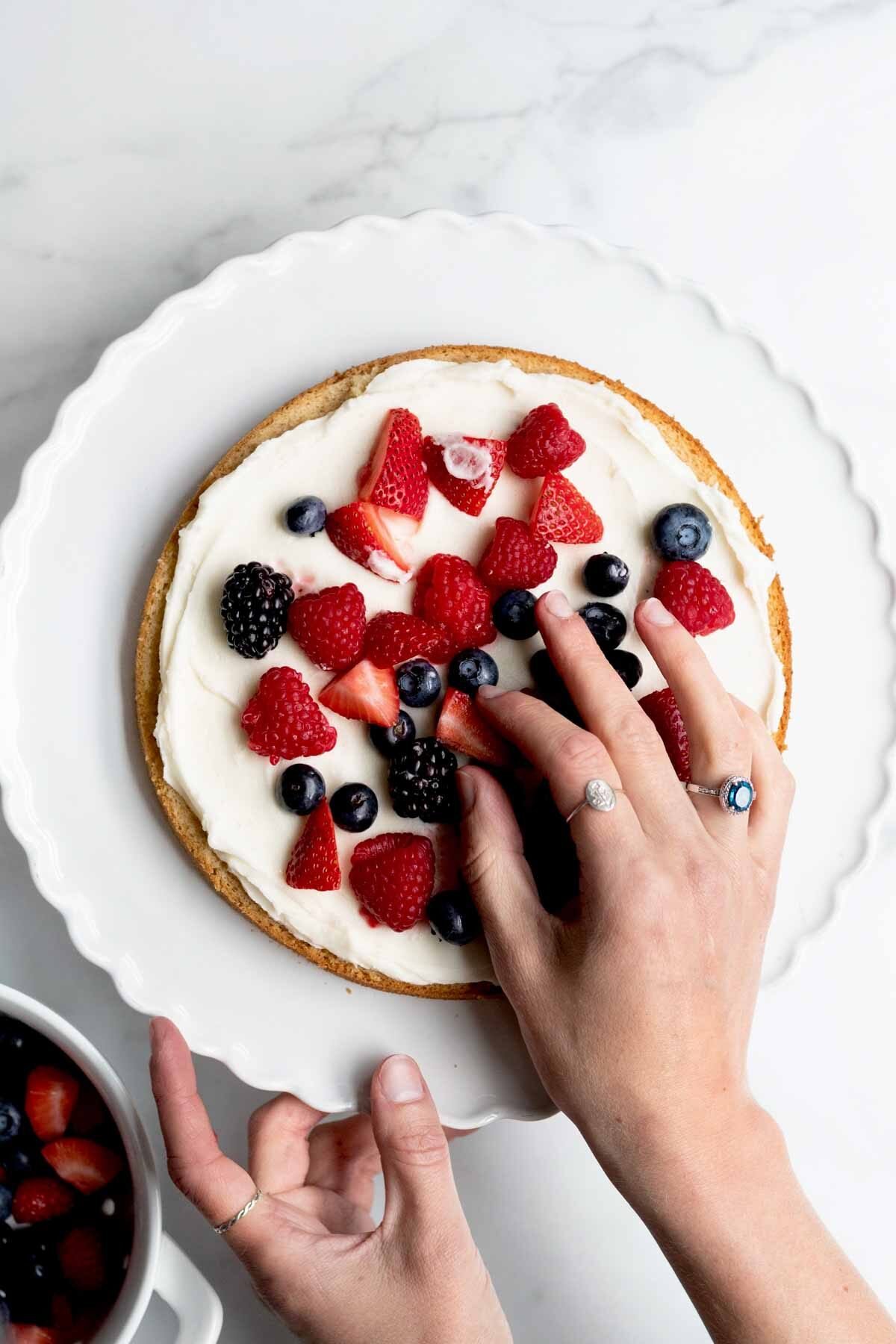 A hand delicately places the last strawberry atop the vanilla frosted cake.