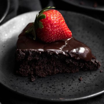 A delicious slice of vegan flourless chocolate cake with a strawberry on top on a black plate.