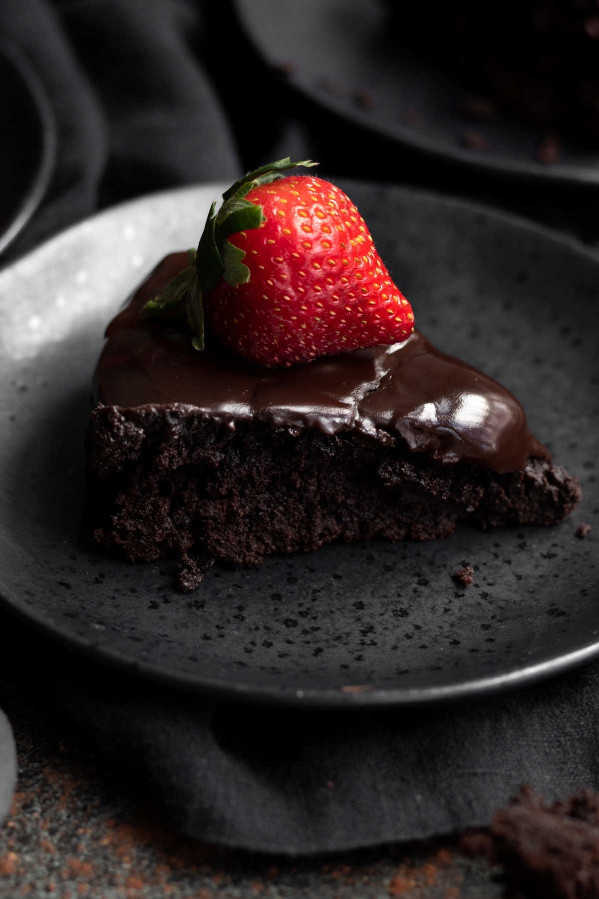 A delicious slice of vegan flourless chocolate cake with a strawberry on top on a black plate.