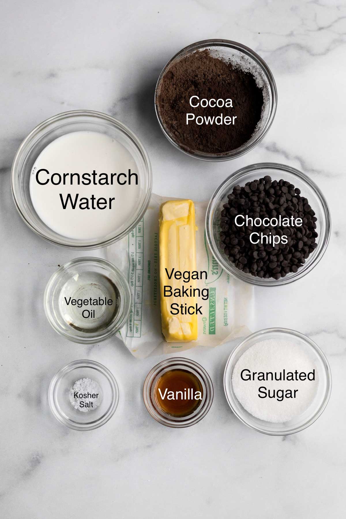 Cornstarch water, cocoa powder, chocolate chips, vegetable oil, a vegan baking stick, kosher salt, vanilla and granulated sugar in separate glass bowls.