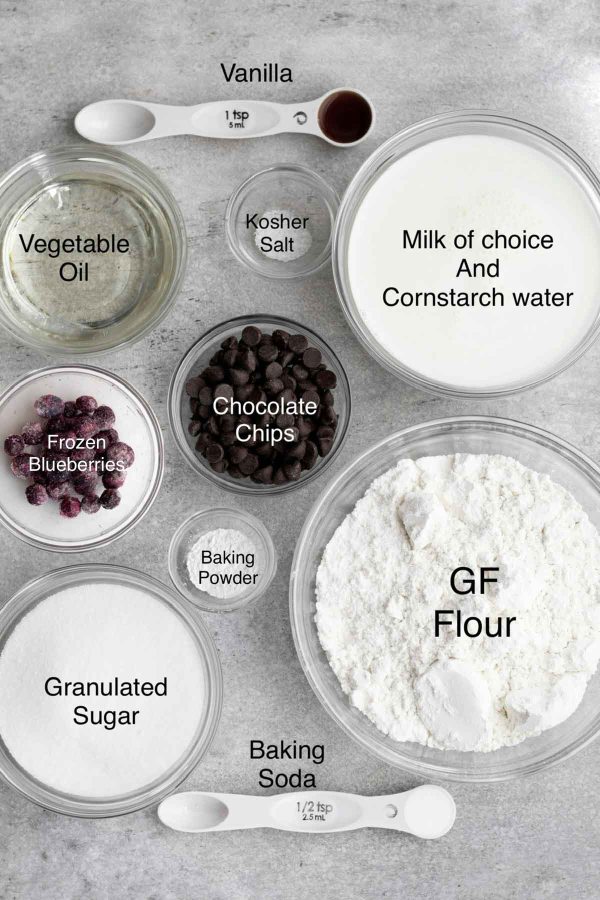 Vanilla, vegetable oil, kosher salt, milk and cornstarch water, frozen blueberries, chocolate chips, baking powder, gluten free flour, granulated sugar and baking soda in separate containers.