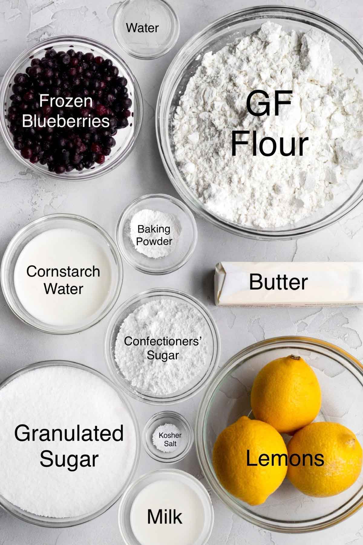 Frozen blueberries, water, gluten free flour, cornstarch water, baking powder, butter, confectioners' sugar, granulated sugar, kosher salt, three whole lemons, and milk in separate containers.
