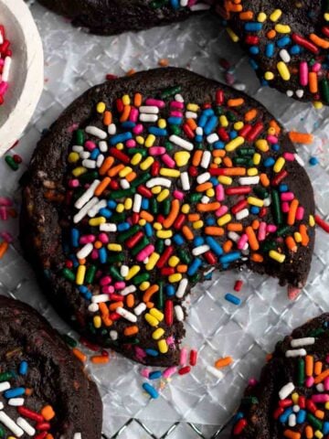 Bright rainbow-colored sprinkles atop a chewy fudgey Chocolate Sprinkle Cookie with a bite taken out.