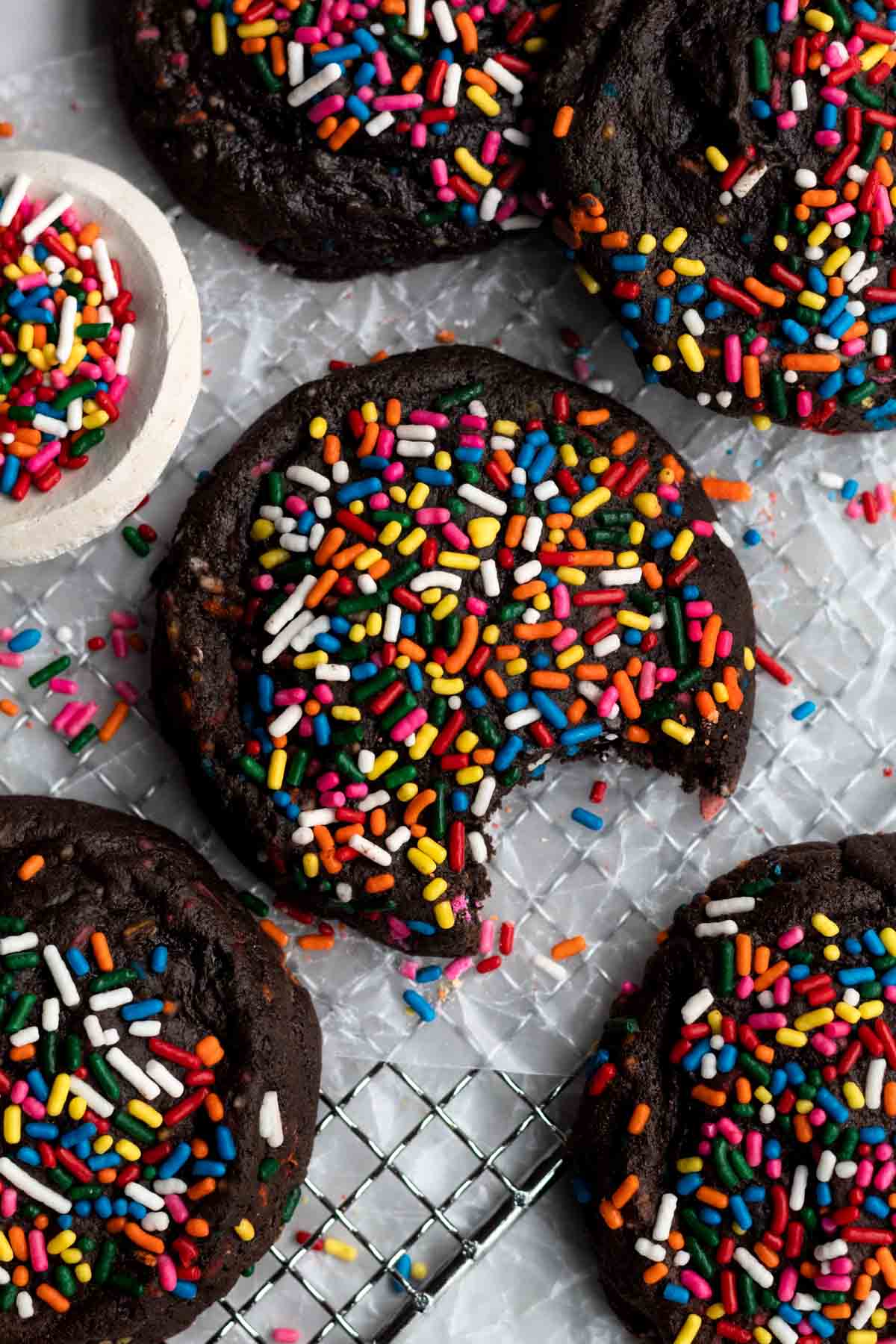 Bright rainbow-colored sprinkles atop a chewy fudgey Chocolate Sprinkle Cookie with a bite taken out.