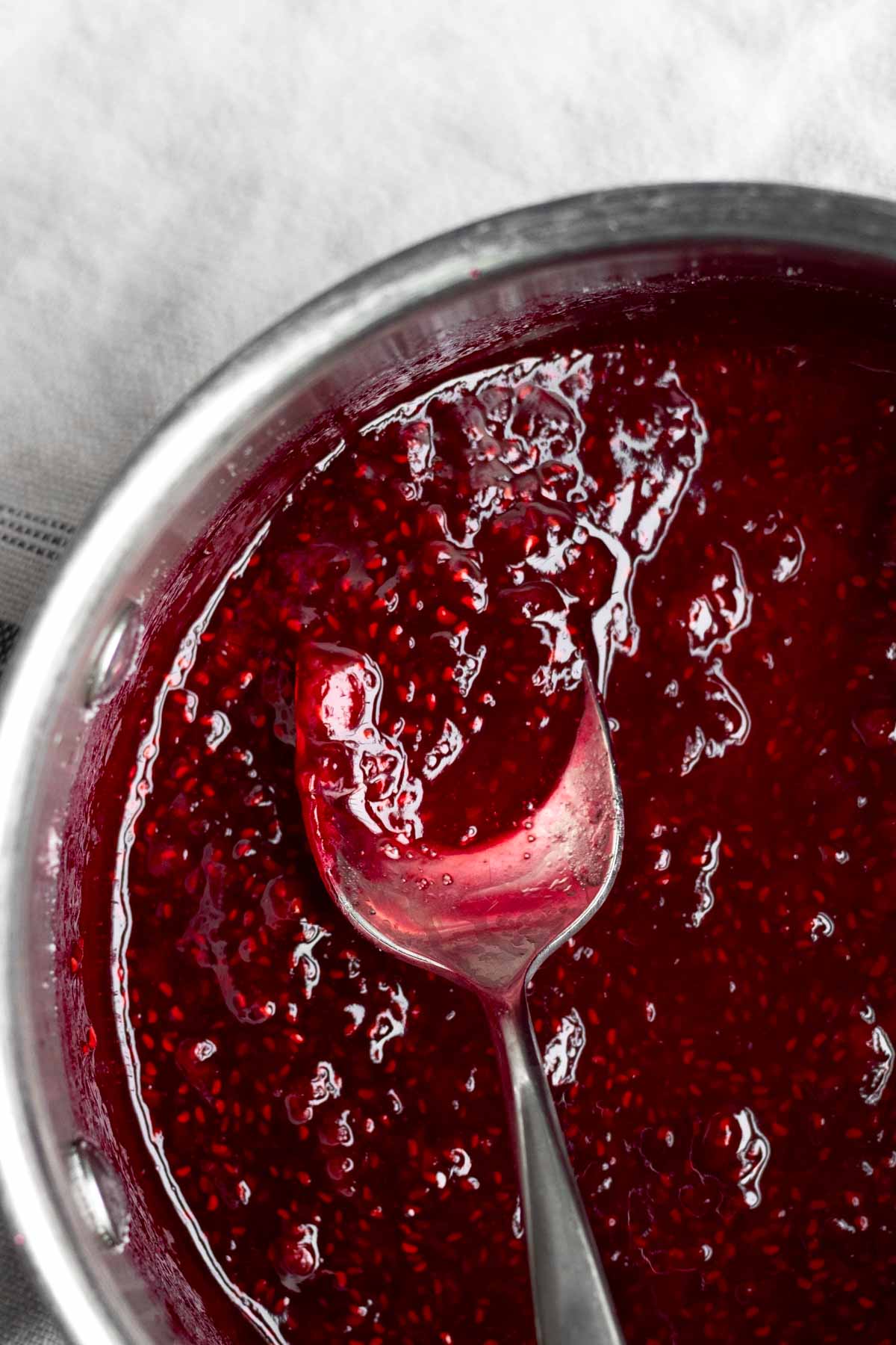 Bright red sugary raspberry sauce in a saucepan with a spoon.