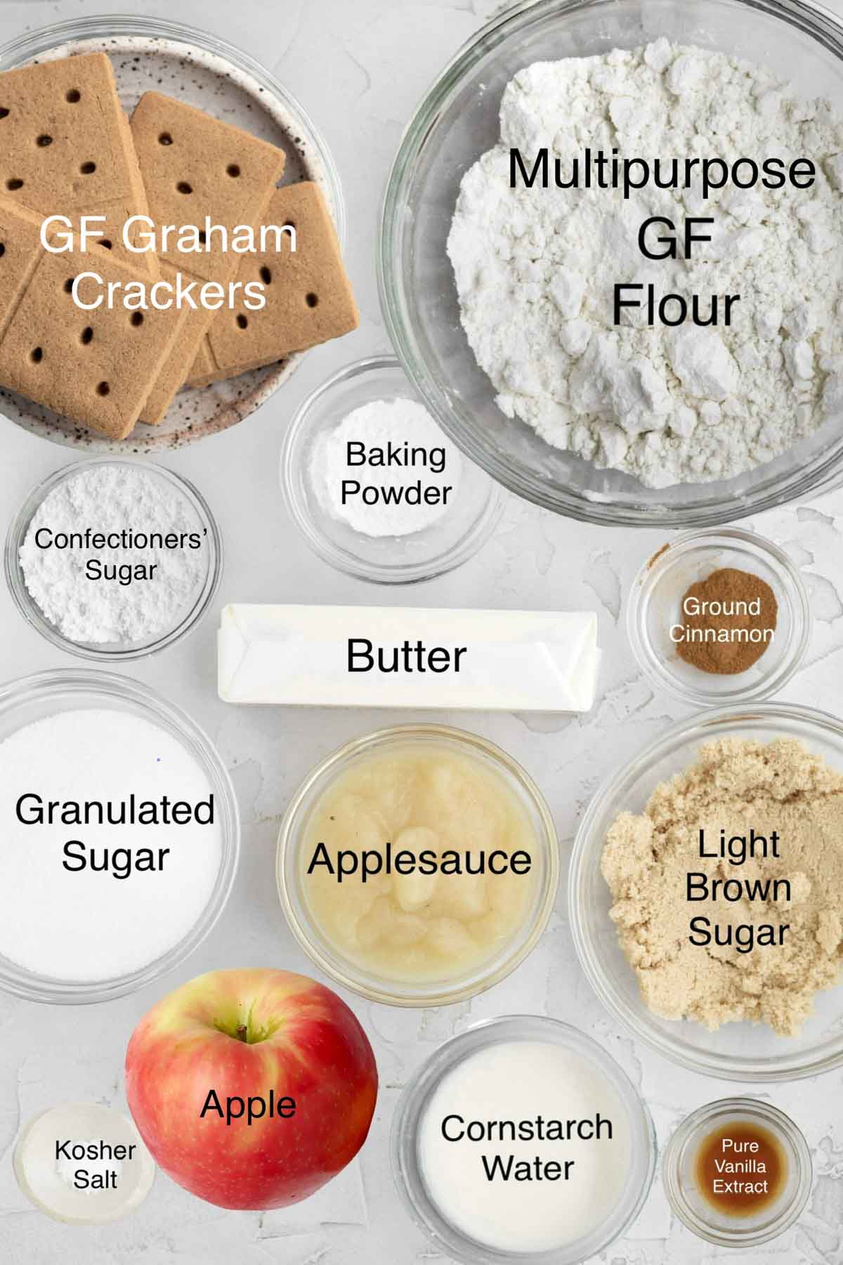 Gluten free graham crackers, multipurpose gluten free flour, confectioners' sugar, baking powder, butter, ground cinnamon, granulated sugar, applesauce, light brown sugar, kosher salt, an apple, cornstarch water, and pure vanilla extract in separate containers.