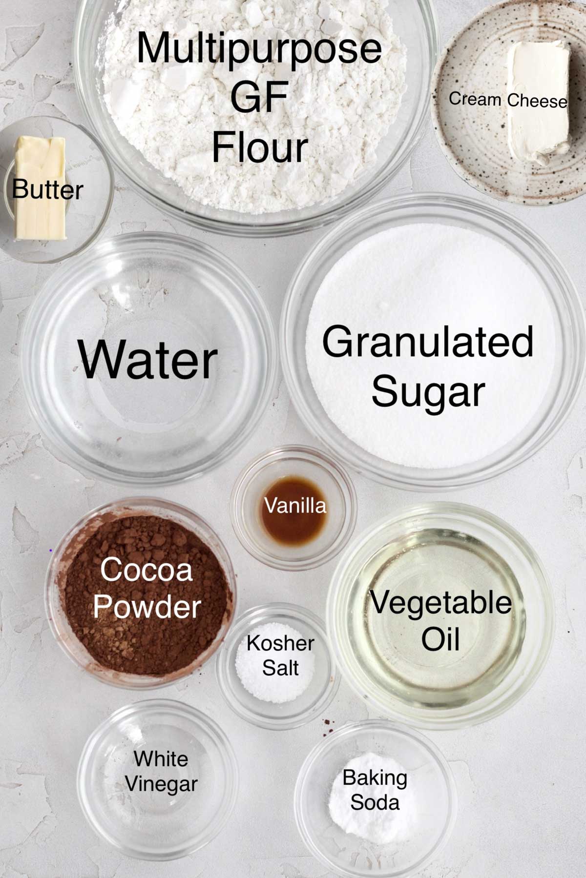 Separate bowls of butter, multipurpose gluten free flour, cream cheese, water, granulated sugar, vanilla, cocoa powder, vegetable oil, kosher salt, white vinegar, and baking soda.