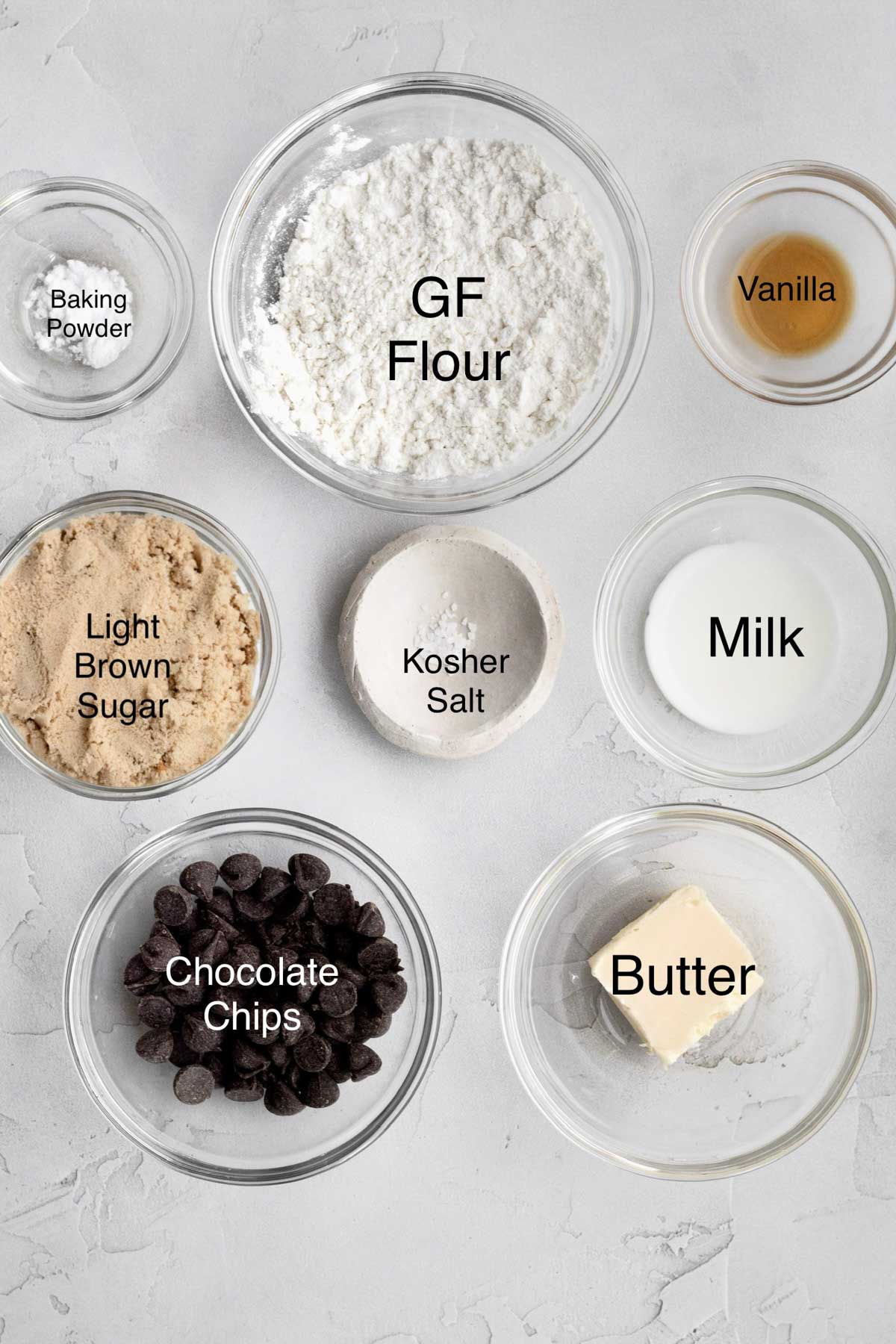 Bowls of baking powder, gluten free flour, vanilla, light brown sugar, kosher salt, milk, chocolate chips and butter.