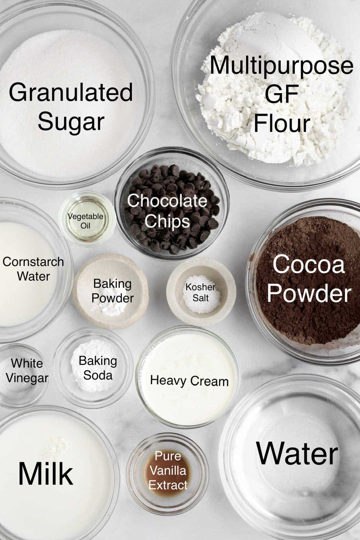 Bowls of granulated sugar, multipurpose gluten free flour, vegetable oil, chocolate chips, cornstarch water, baking powder, kosher salt, cocoa powder, white vinegar, baking soda, heavy cream, milk, pure vanilla extract and water.