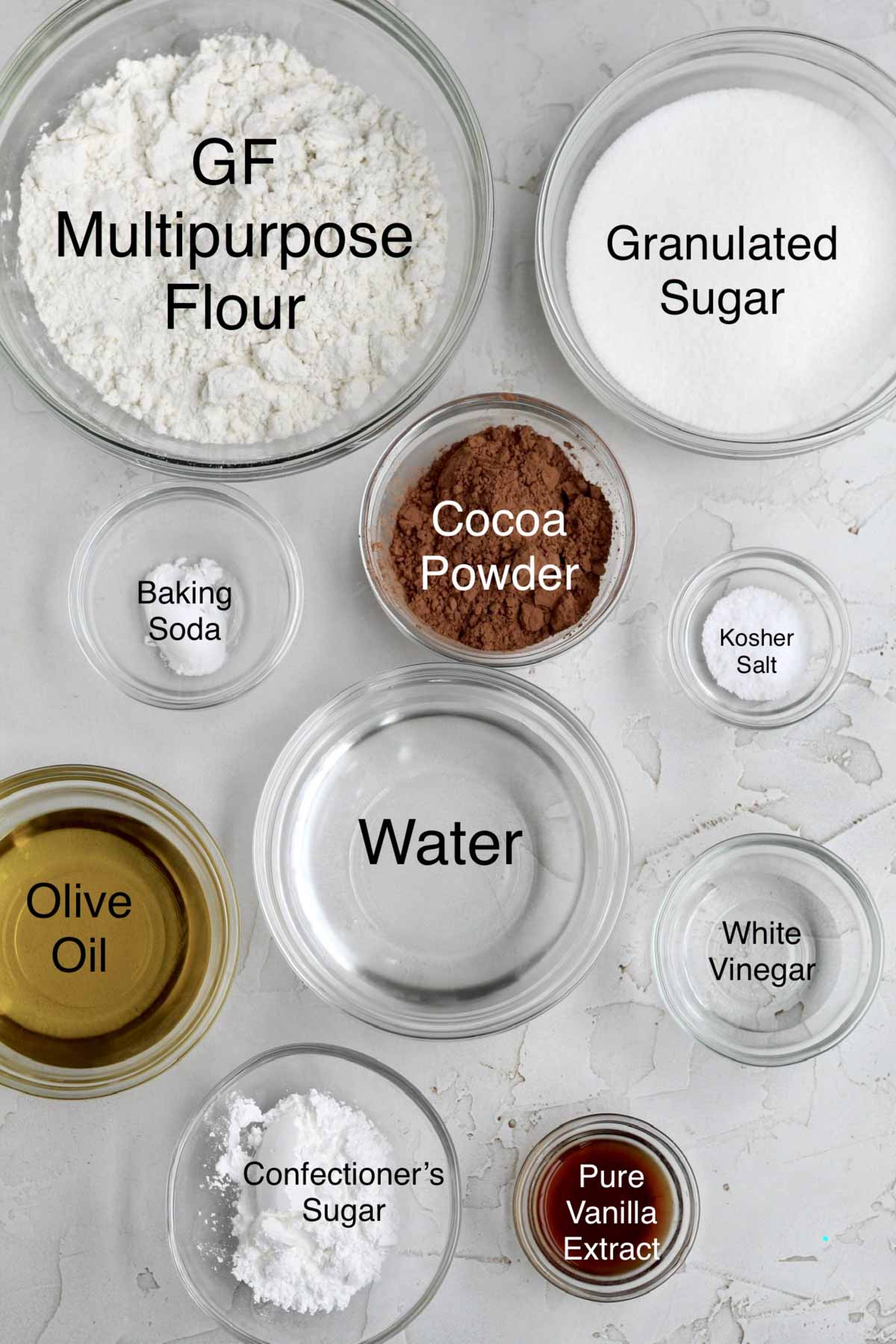 Bowls of gluten free flour, granulated sugar, baking soda, cocoa powder, kosher salt, olive oil, water, white vinegar, confectioners' sugar, and pure vanilla extract.