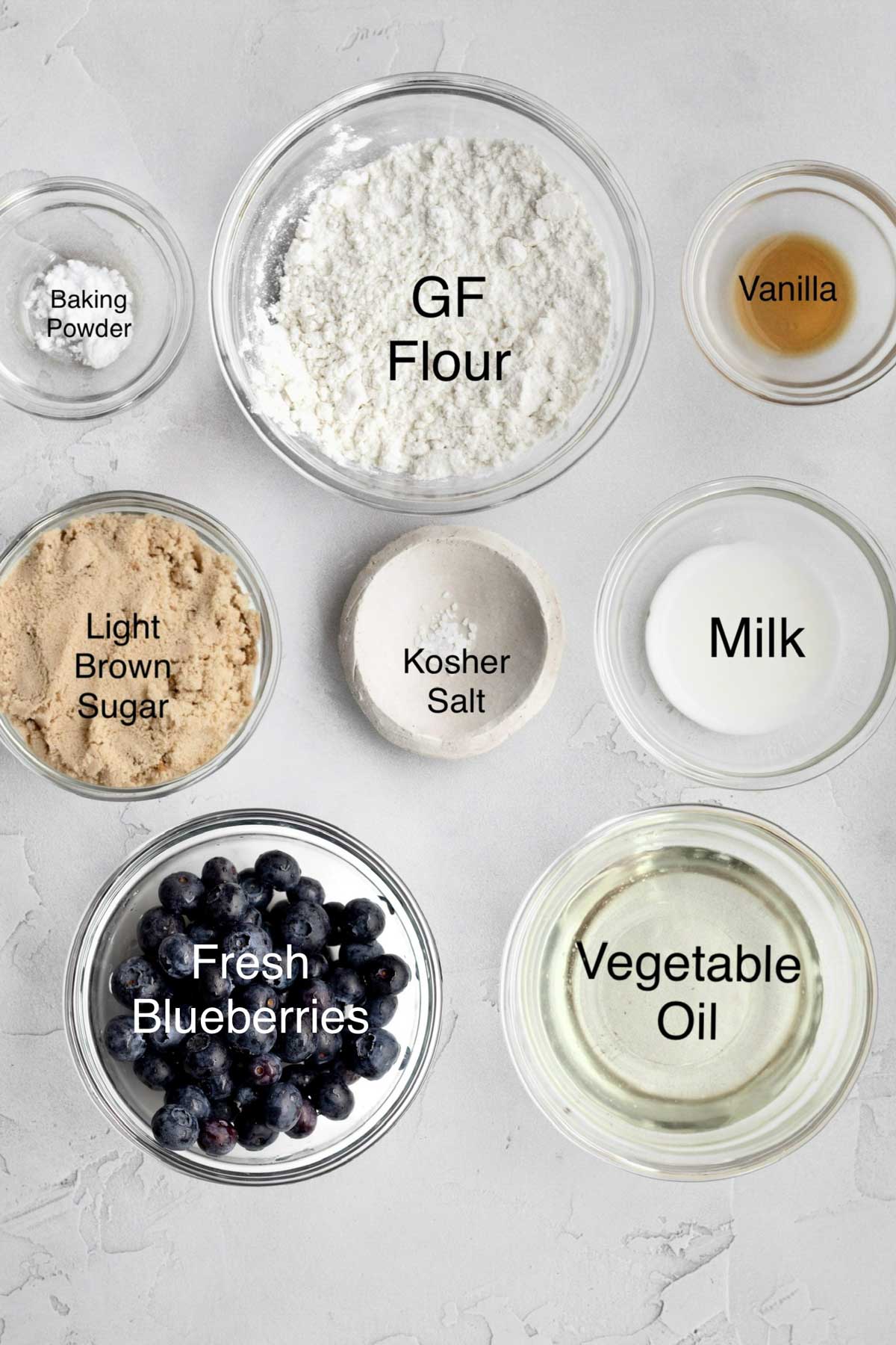 Bowls of baking powder, gluten free flour, vanilla, light brown sugar, kosher salt, milk, fresh blueberries and vegetable oil.