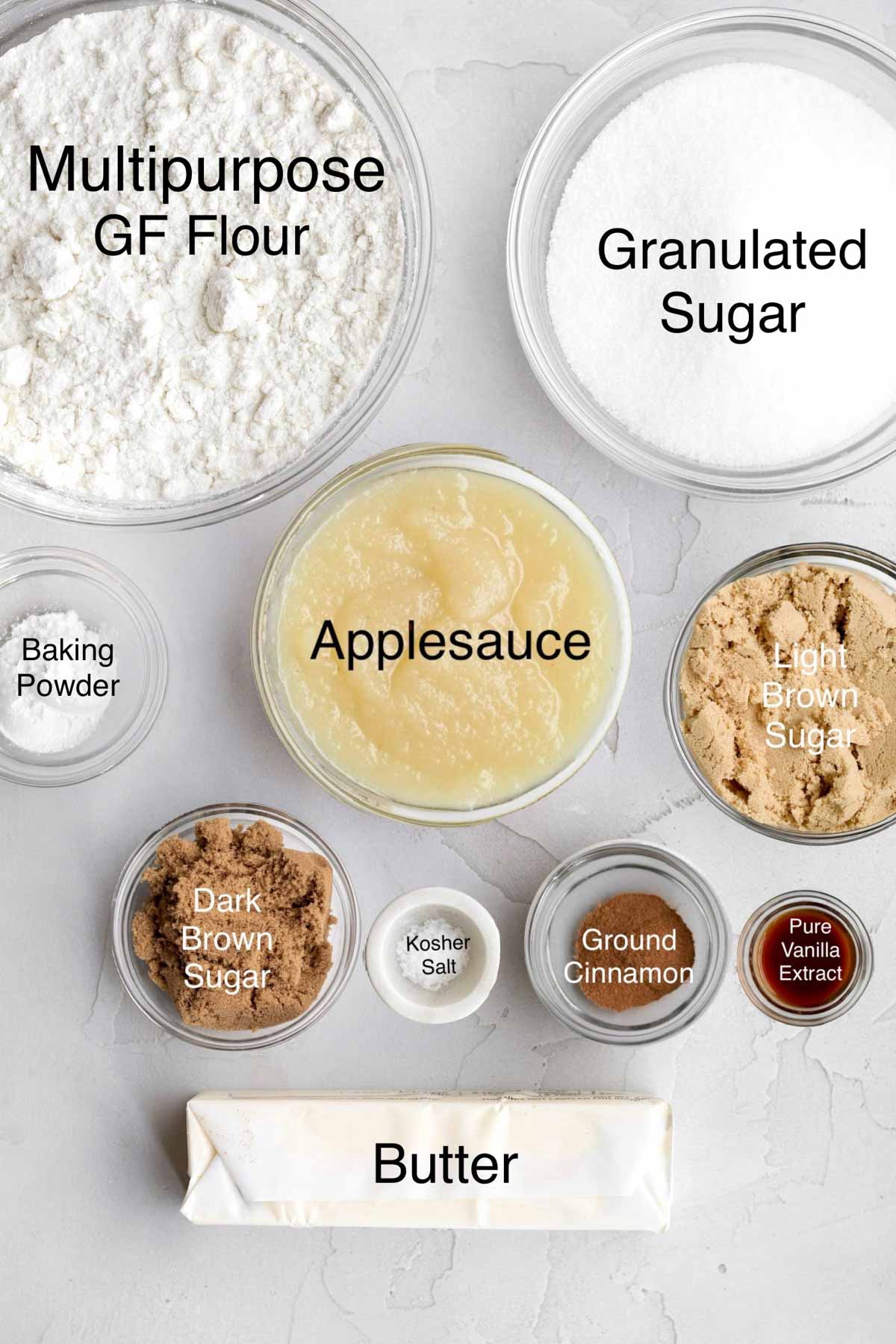 Separate containers of Gluten Free Flour, granulated sugar, baking powder, applesauce, light brown sugar, dark brown sugar, kosher salt, ground cinnamon, pure vanilla extract, and butter.