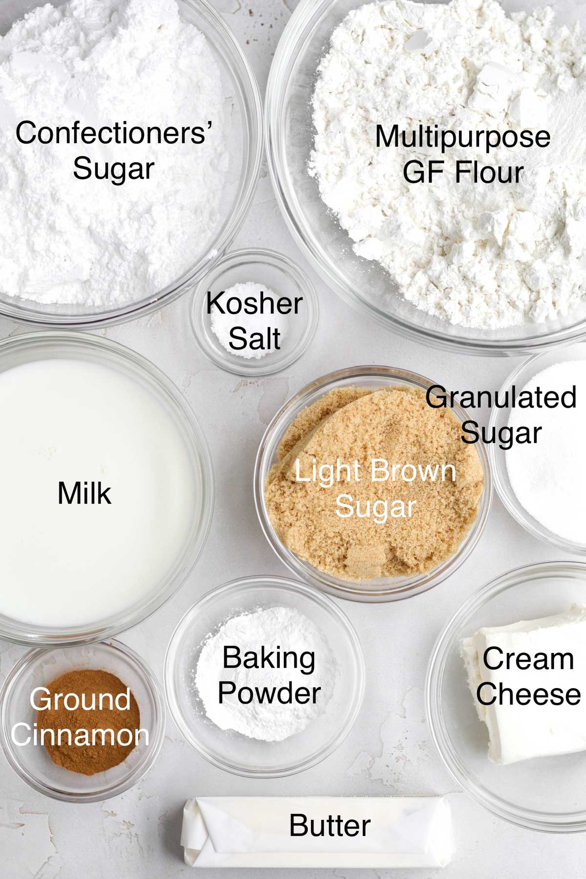 Bowls of confectioners' sugar, multipurpose gluten free flour, kosher salt, milk, light brown sugar, granulated sugar, ground cinnamon, baking powder, cream cheese, and butter.