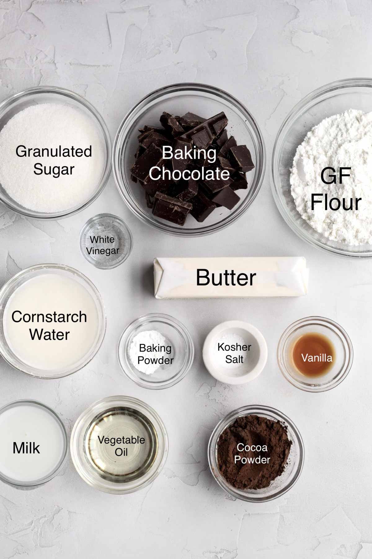 Bowls of granulated sugar, baking chocolate, gluten free flour, white vinegar, butter, cornstarch water, baking powder, kosher salt, vanilla, milk, vegetable oil, and cocoa powder.