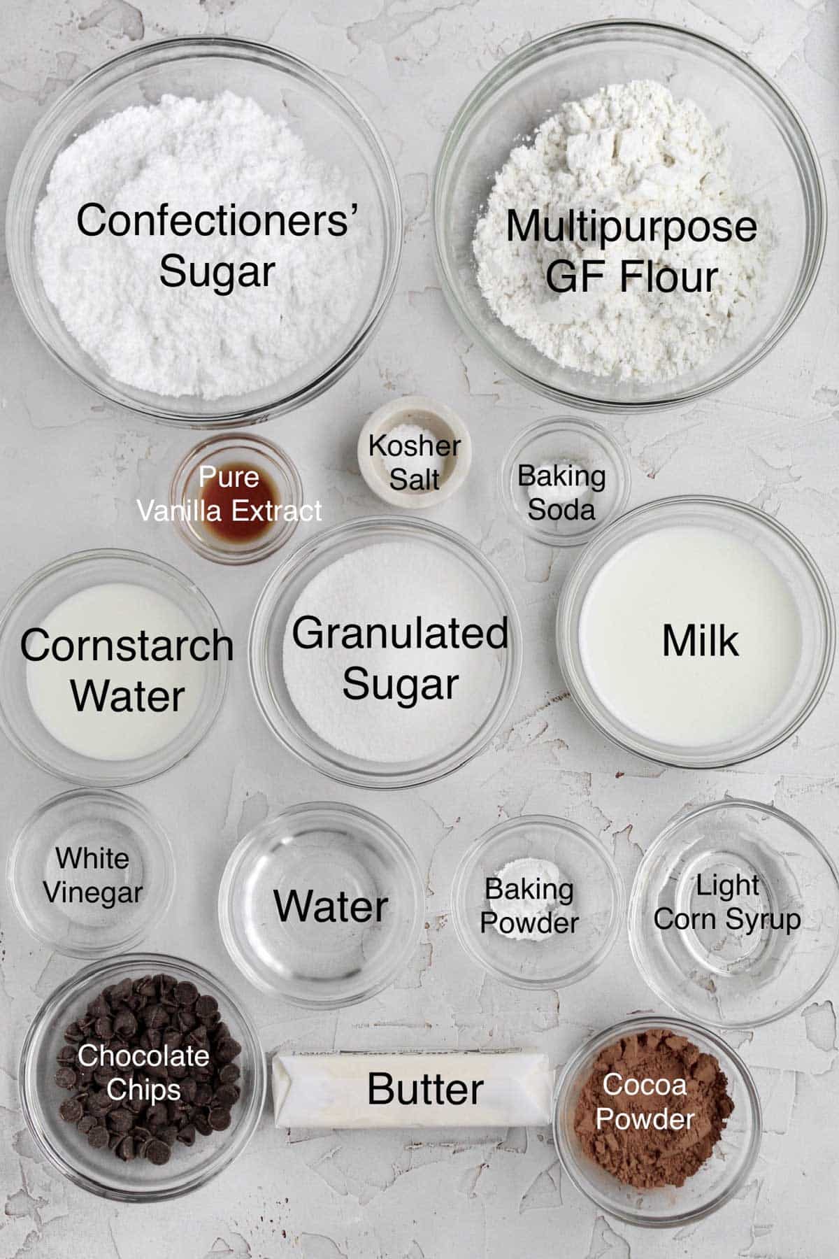 Bowls of confectioners' sugar, multipurpose gluten free flour, pure vanilla extract, kosher salt, baking soda, cornstarch water, granulated sugar, milk, white vinegar, water, baking powder, light corn syrup, chocolate chips, butter and cocoa powder.