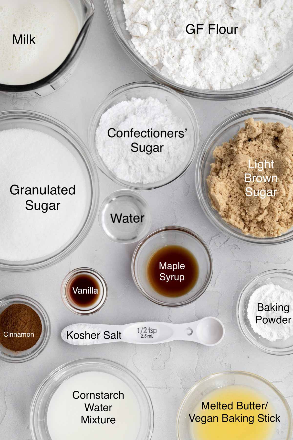 Separate bowls of milk, gluten free flour, granulated sugar, confectioners' sugar, light brown sugar, water, cinnamon, vanilla, maple syrup, baking powder, kosher salt, cornstarch water, and melted butter.