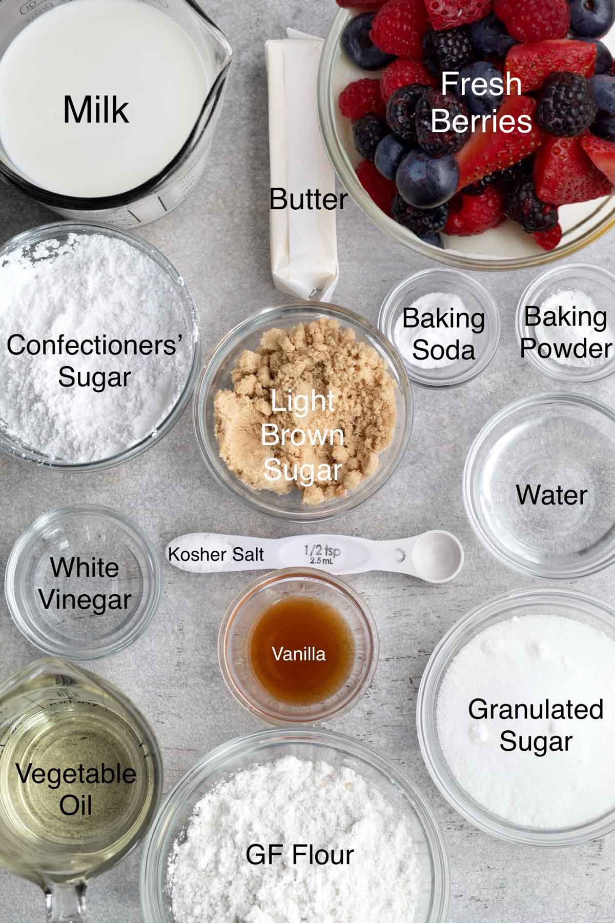 mixed berry cake ingredients in separate containers: milk, butter, fresh berries, confectioners' sugar, light brown sugar, baking soda, baking powder, white vinegar, kosher salt, water, vanilla, vegetable oil, gluten free flour, and granulated sugar.
