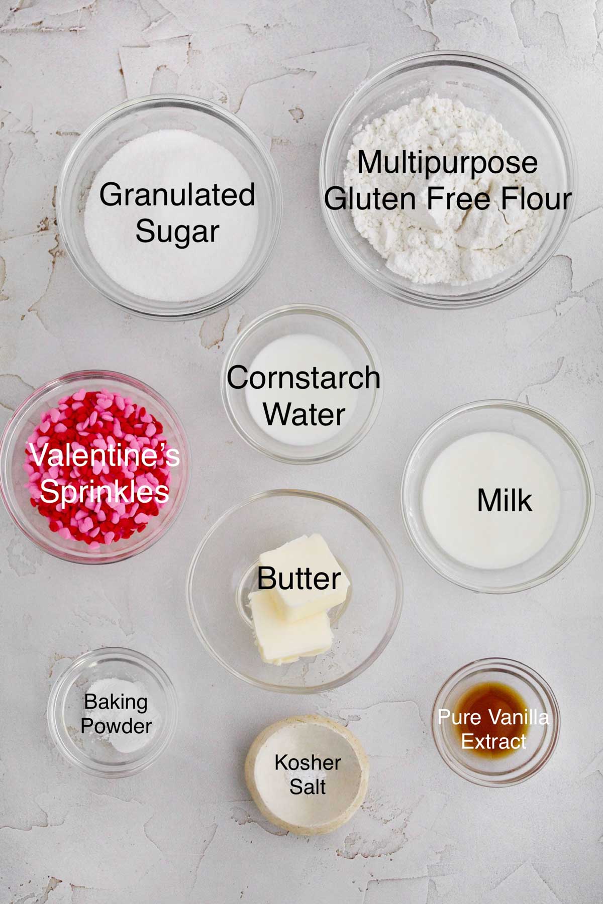 Bowls of granulated sugar, multipurpose gluten free flour, Valentine's sprinkles, cornstarch water, butter, milk, baking powder, kosher salt, and pure vanilla extract.