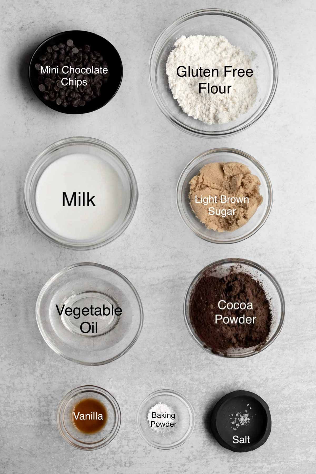 easy chocolate mug cakes ingredients in separate bowls: mini chocolate chips, gluten free flour, milk, light brown sugar, vegetable oil, cocoa powder, vanilla, baking powder, and salt.
