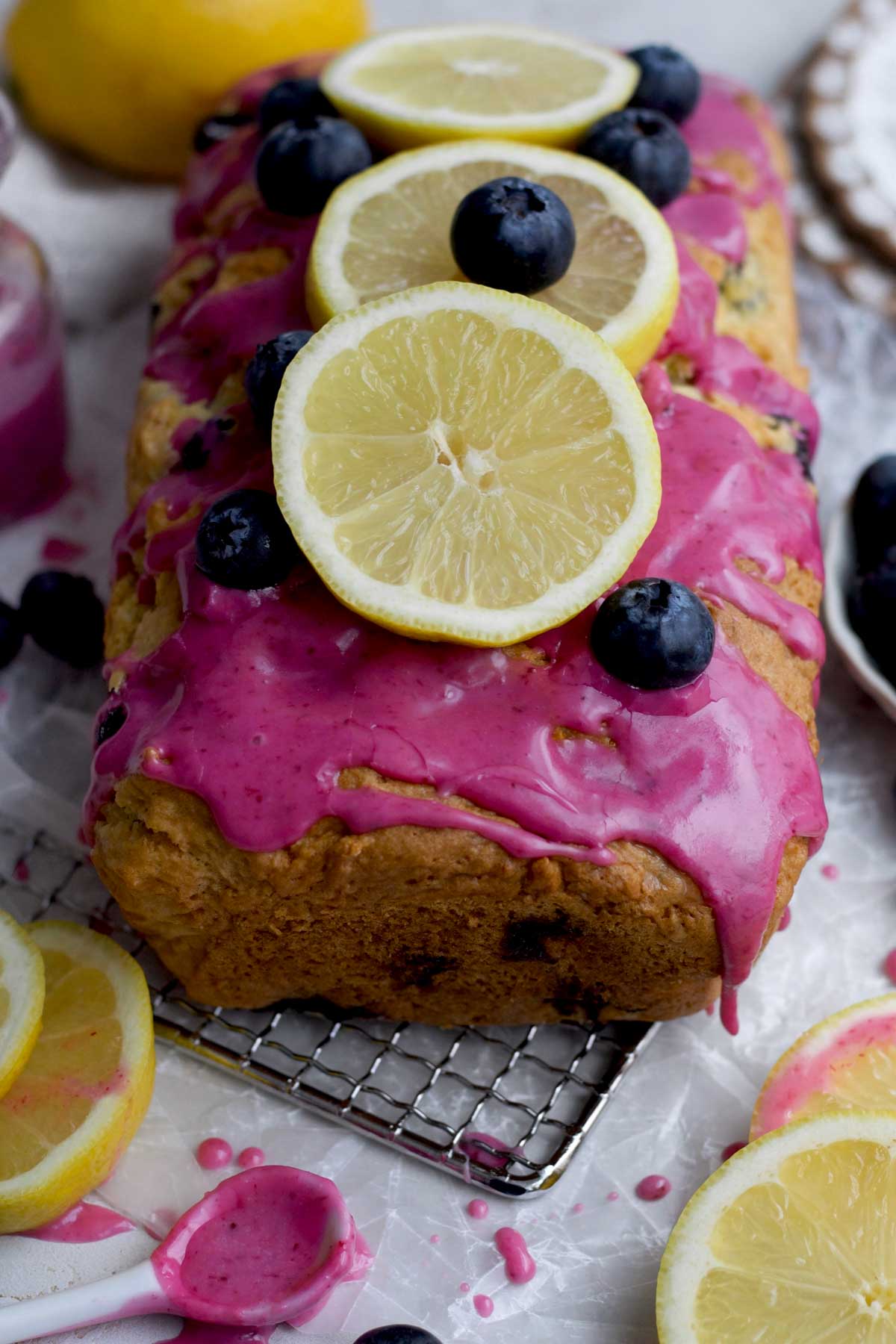 Thick vibrant blueberry glaze drips down the golden brown eggless lemon blueberry loaf topped with lemons.