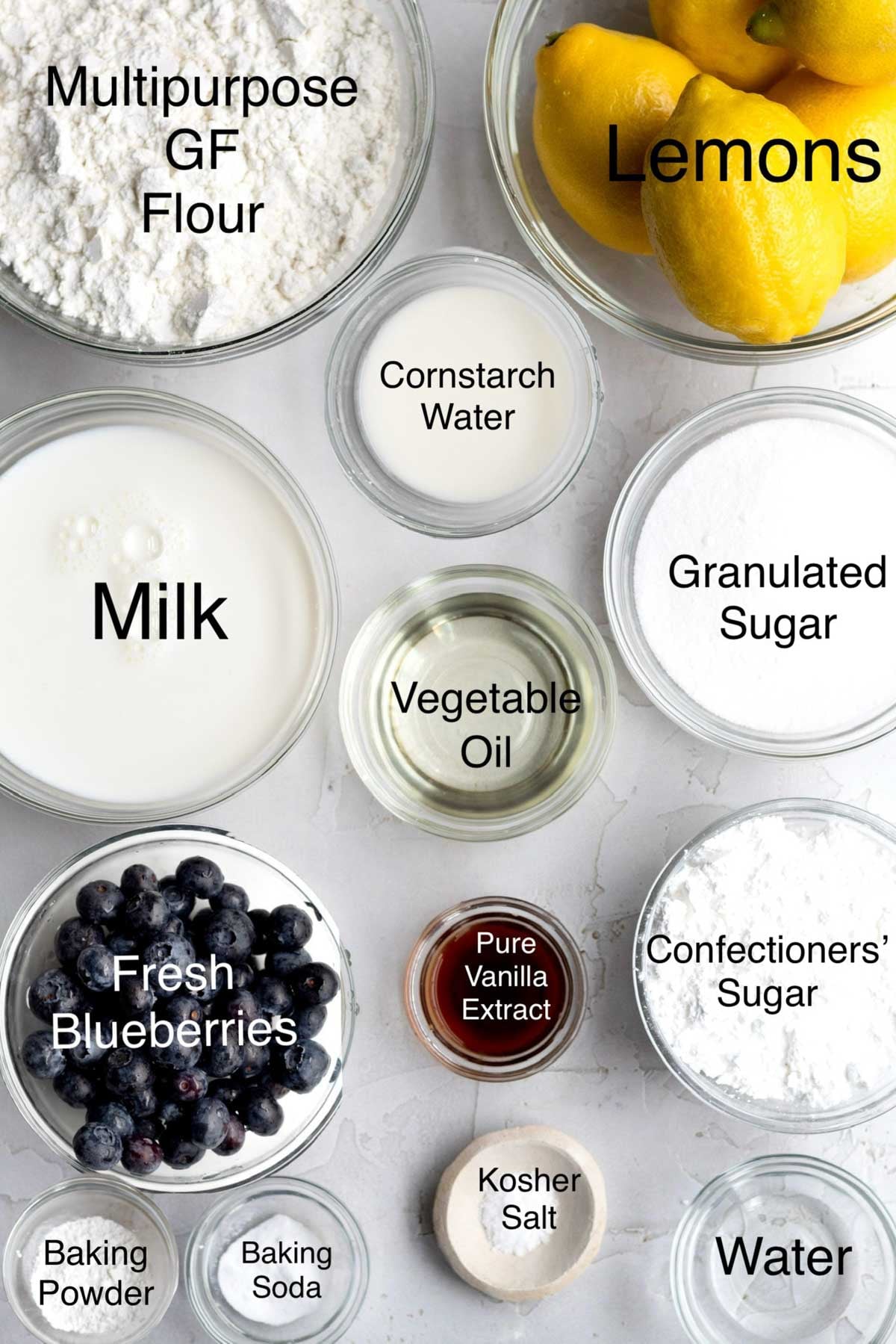 Lemon Blueberry loaf ingredients in separate bowls: multipurpose gluten free flour, lemons, cornstarch water, milk, vegetable oil, granulated sugar, fresh blueberries, pure vanilla extract, confectioners' sugar, baking powder, baking soda, kosher salt, and water.