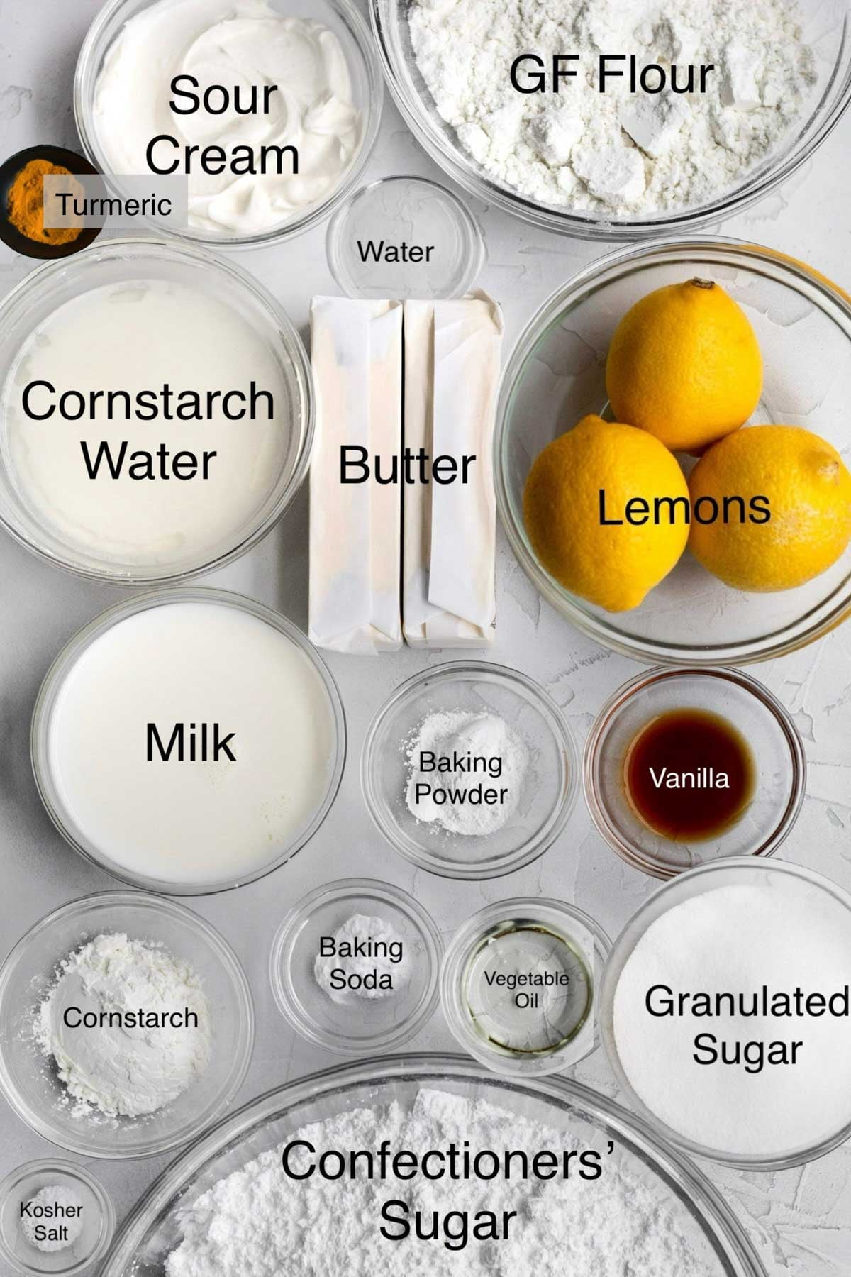 Lemon curd cupcakes ingredients in separate containers: turmeric, sour cream, gluten free flour, cornstarch water, water, butter, lemons, milk, baking powder, vanilla, cornstarch, baking soda, vegetable oil, granulated sugar, kosher salt, and confectioners' sugar.