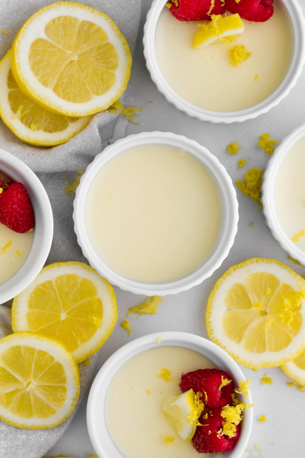 several gluten-free lemon posset desserts in ruffled white ramekins surrounded by lemon slices and zest.