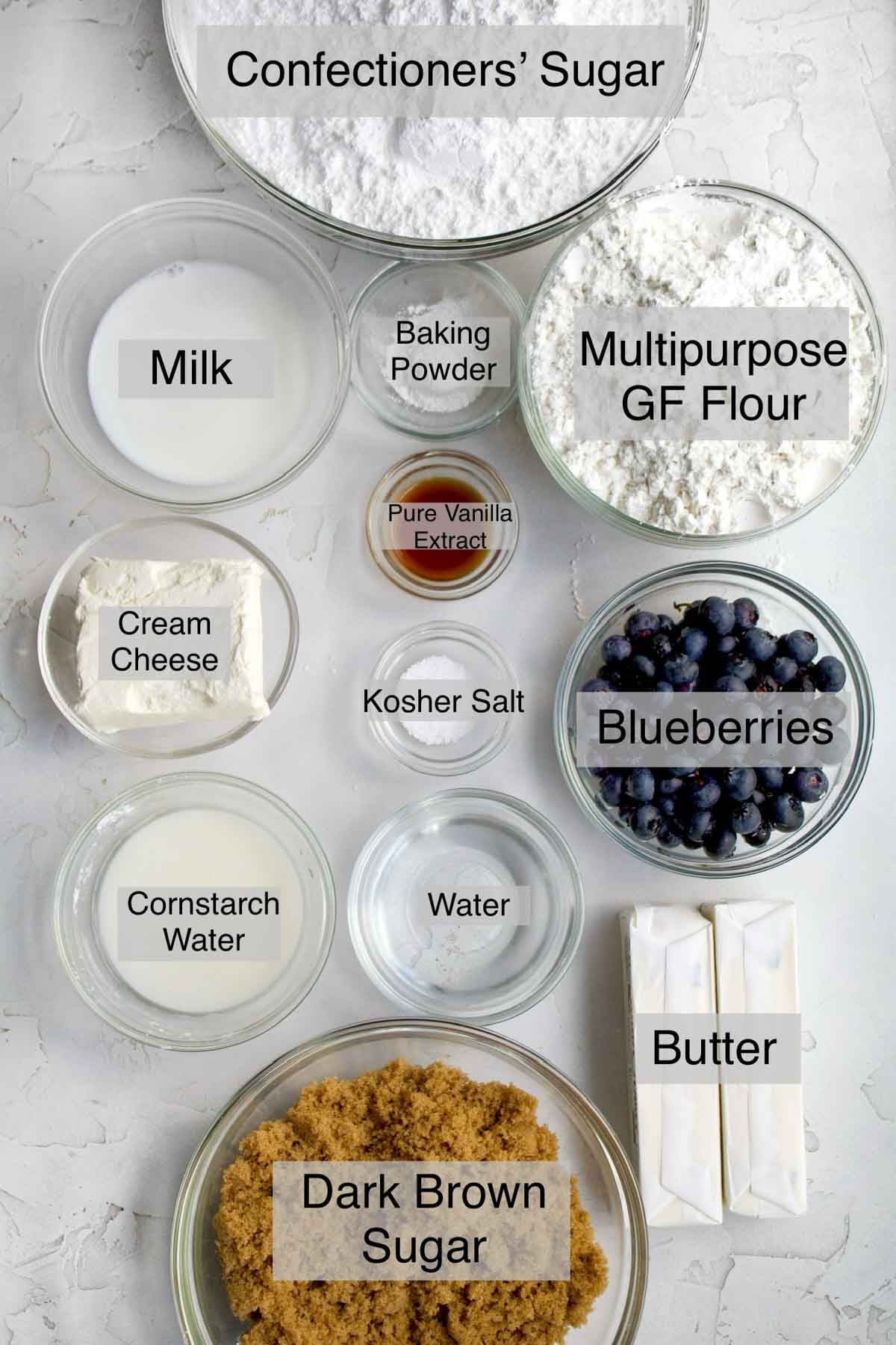brown sugar cookies with blueberry cream cheese frosting ingredients in separate bowls: confectioners' sugar, milk, baking powder, multipurpose gluten free flour, pure vanilla extract, cream cheese, kosher salt, blueberries, cornstarch water, butter and dark brown sugar.