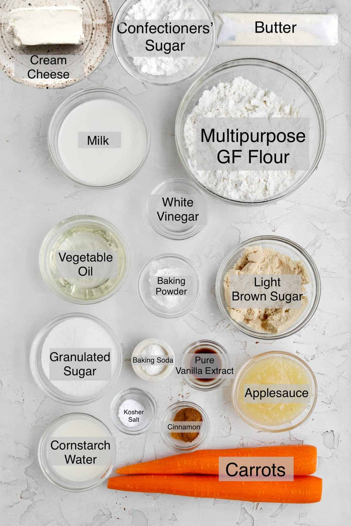 easy carrot cake ingredients in sepate containers: cream cheese, confectioners' sugar, butter, milk, multipurpose gluten free flour, white vinegar, vegetable oil, baking powder, light brown sugar, granulated sugar, baking soda, pure vanilla extract, applesauce, cornstarch water, kosher salt, cinnamon and carrots.