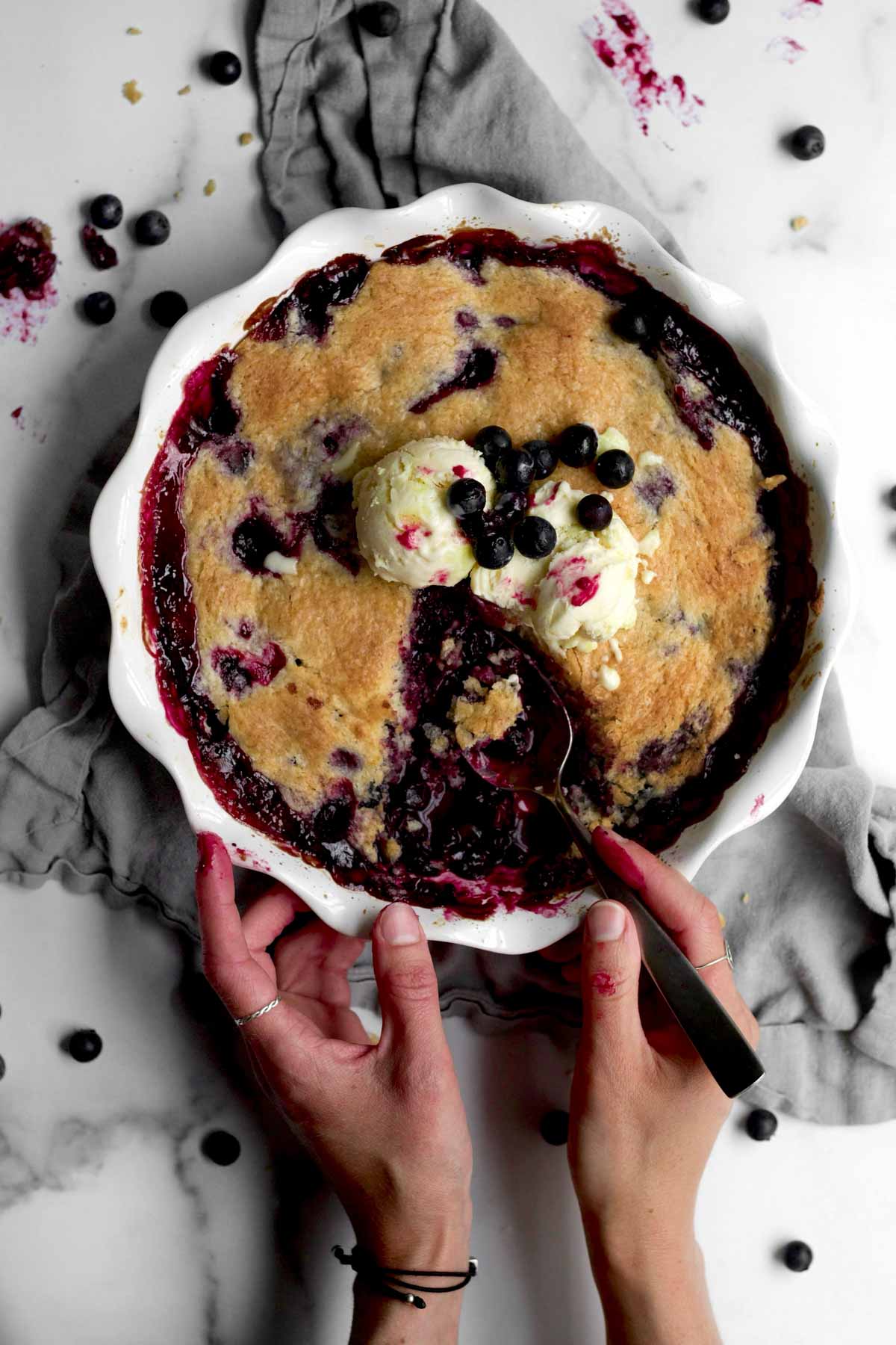 scooping the warm blueberries from the interior of a gluten-free blueberry cobbler topped with ice cream.