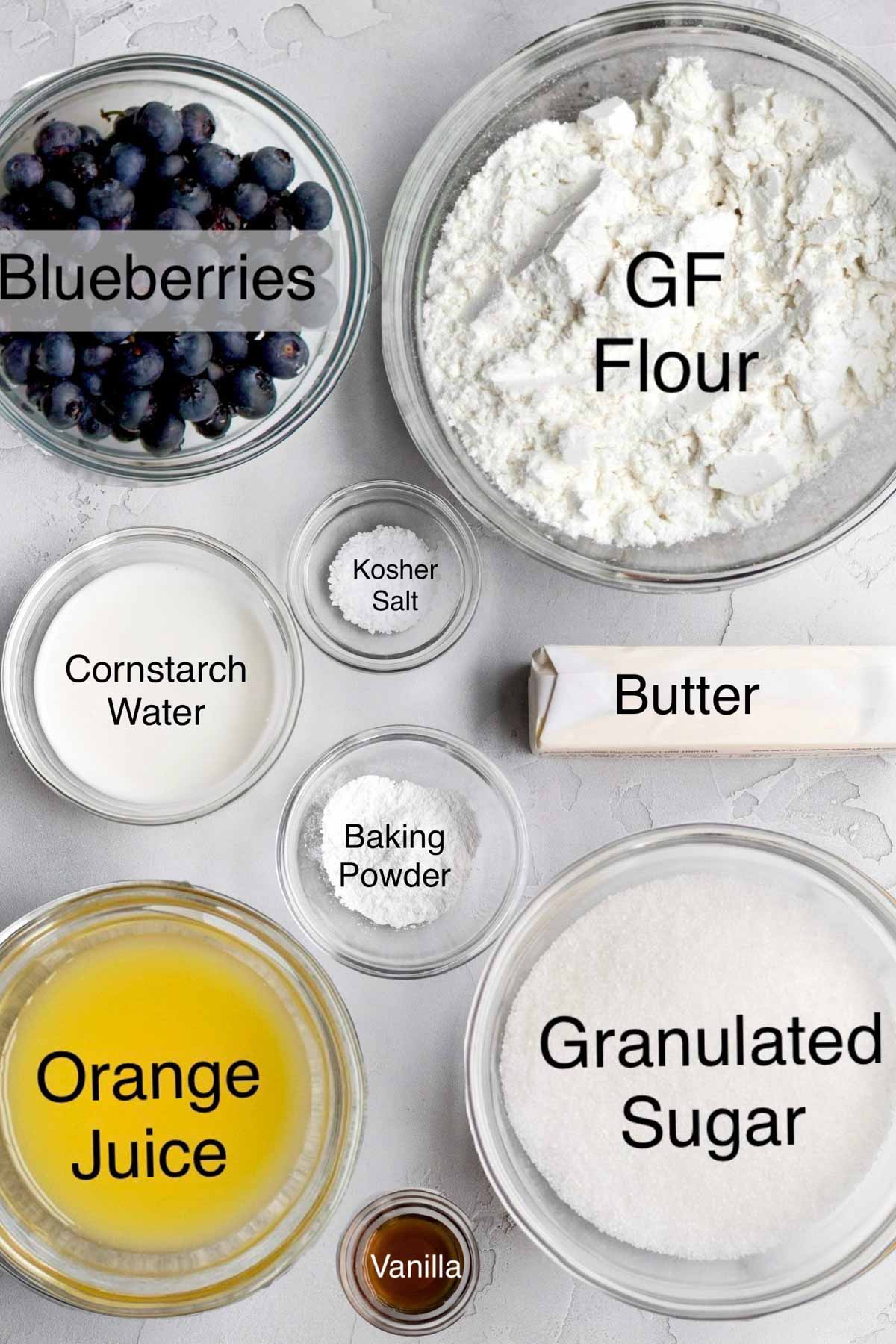 gluten free blueberry cobbler ingredients in separate bowls: blueberries, gluten free flour, cornstarch water, kosher salt, butter, baking powder, orange juice, granulated sugar, and vanilla.
