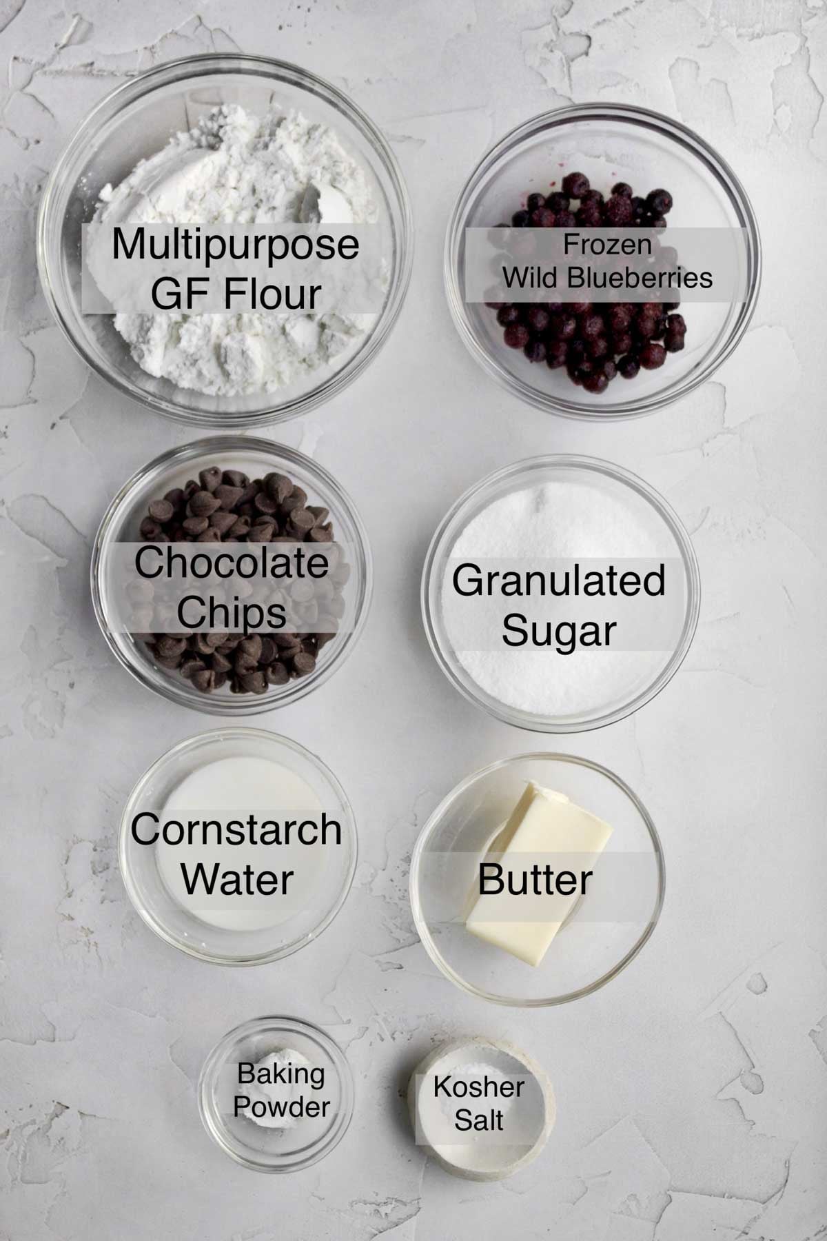Chocolate chip blueberry cookies ingredients in separate bowls: multipurpose gluten fre flour, frozen wild blueberries, chocolate chips, granulated sugar, cornstarch water, butter, baking powder, and kosher salt.