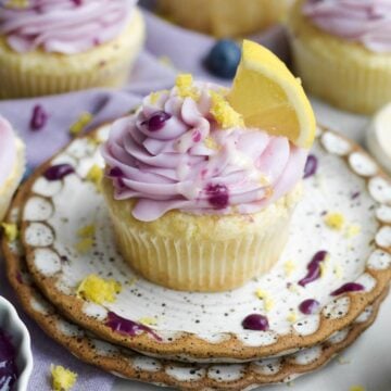 A golden lemon blueberry cupcake with a creamy swirl of lavender blueberry frosting and lemon glaze drizzle.