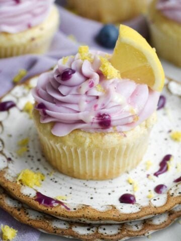 A golden lemon blueberry cupcake with a creamy swirl of lavender blueberry frosting and lemon glaze drizzle.