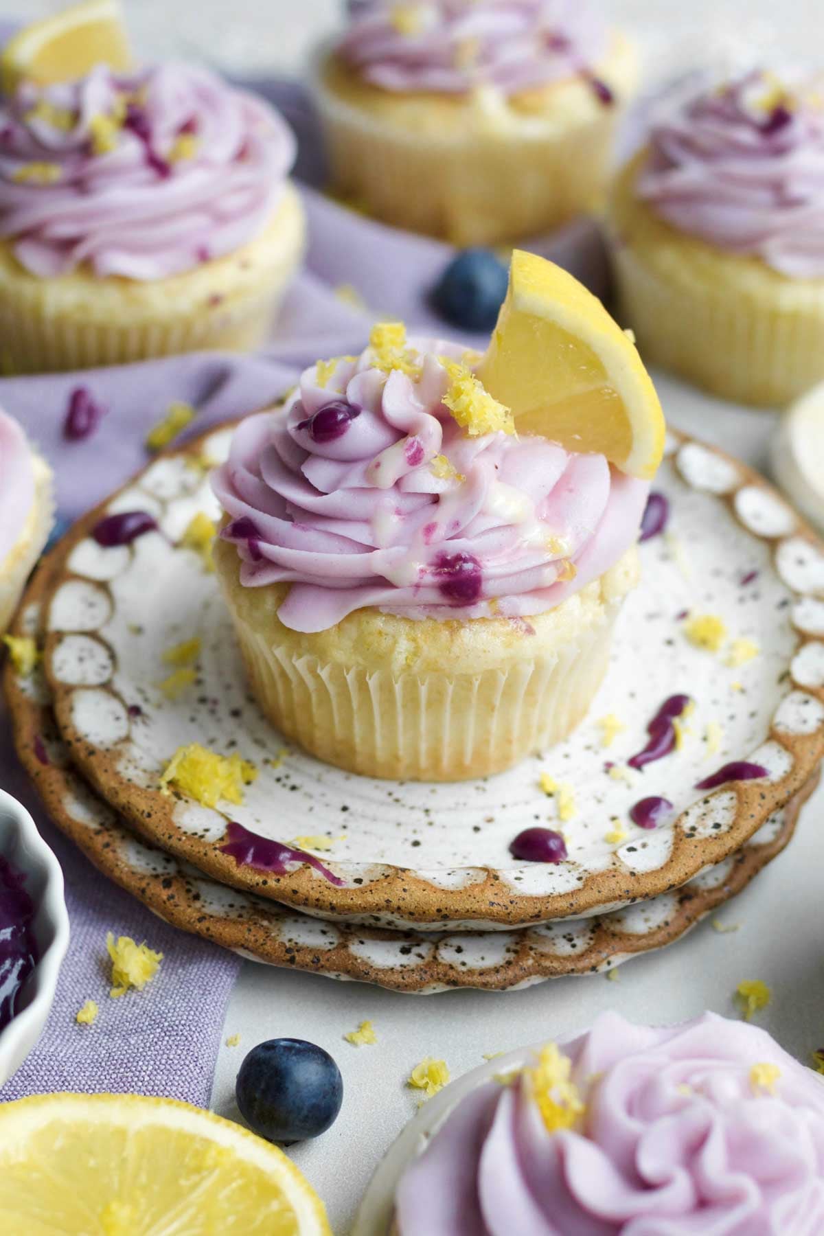 A golden lemon blueberry cupcake with a creamy swirl of lavender blueberry frosting and lemon glaze drizzle.