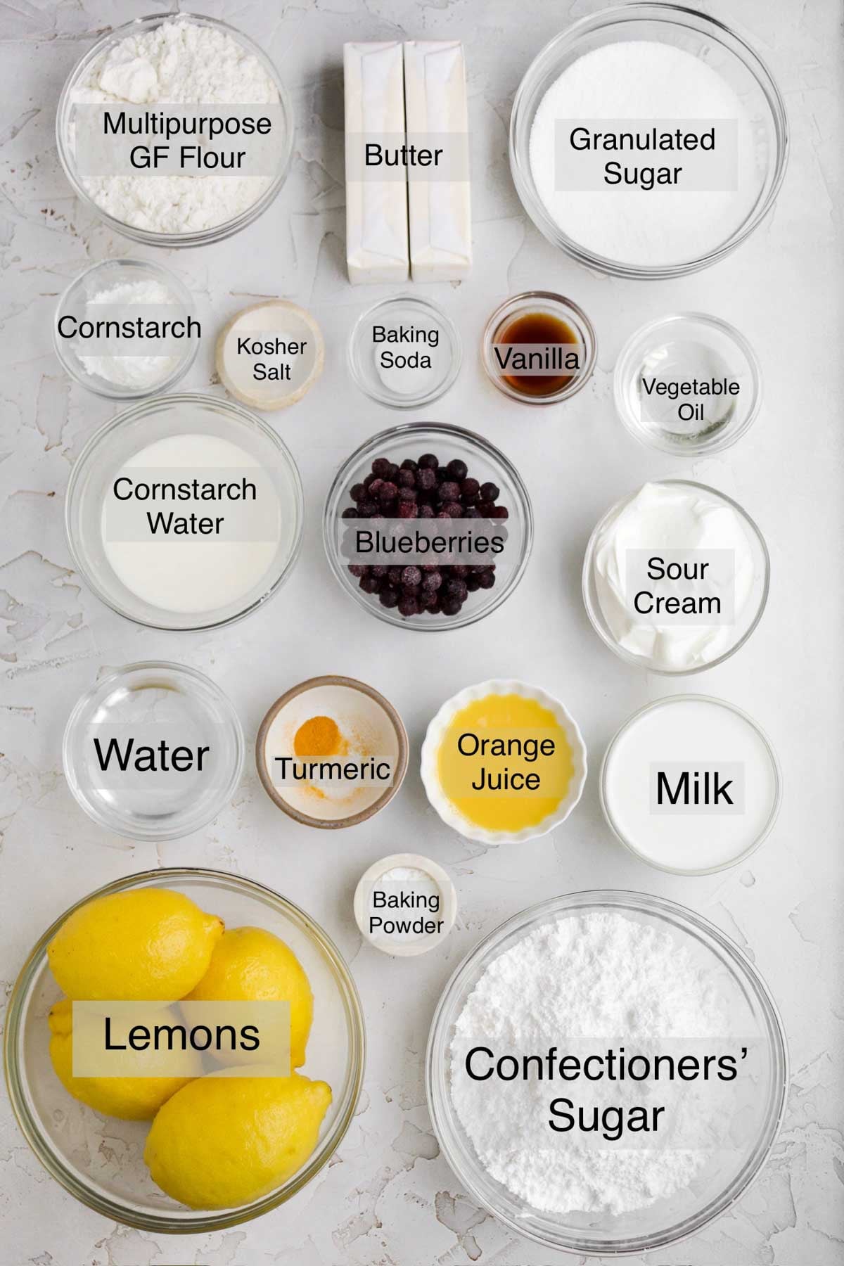 Lemon blueberry cupcakes ingredients in separate containers: multipurpose gluten free flour, butter, granulated sugar, cornstarch, kosher salt, baking soda, vanilla, vegetable oil, cornstarch water, blueberries, sour cream, water, turmeric, orange juice, milk, lemons, baking powder, and confectioners' sugar.