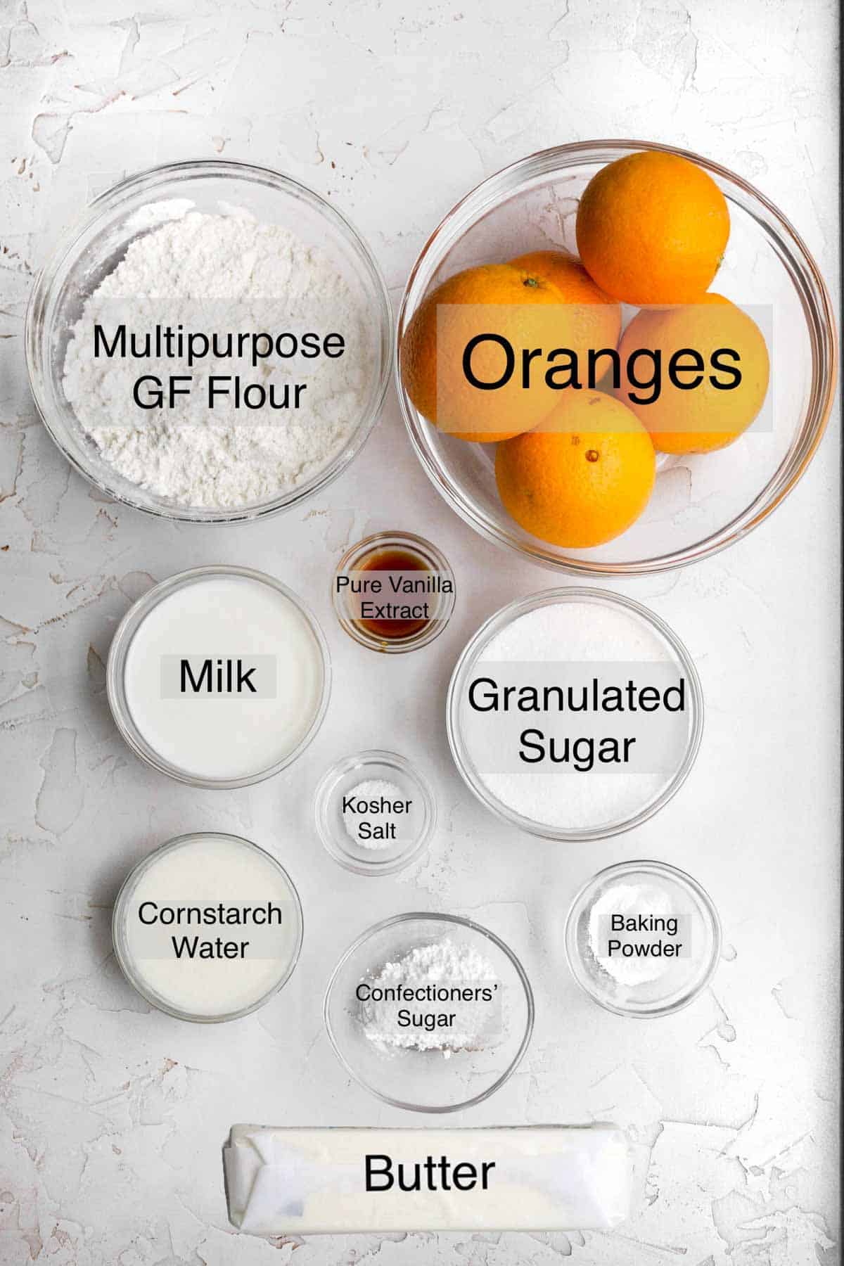 orange cake ingredients in separate bowls: multipurpose gluten free flour, oranges, milk, pure vanilla extract, granulated sugar, kosher salt, cornstarch water, confectioners' sugar, baking powder, and butter.