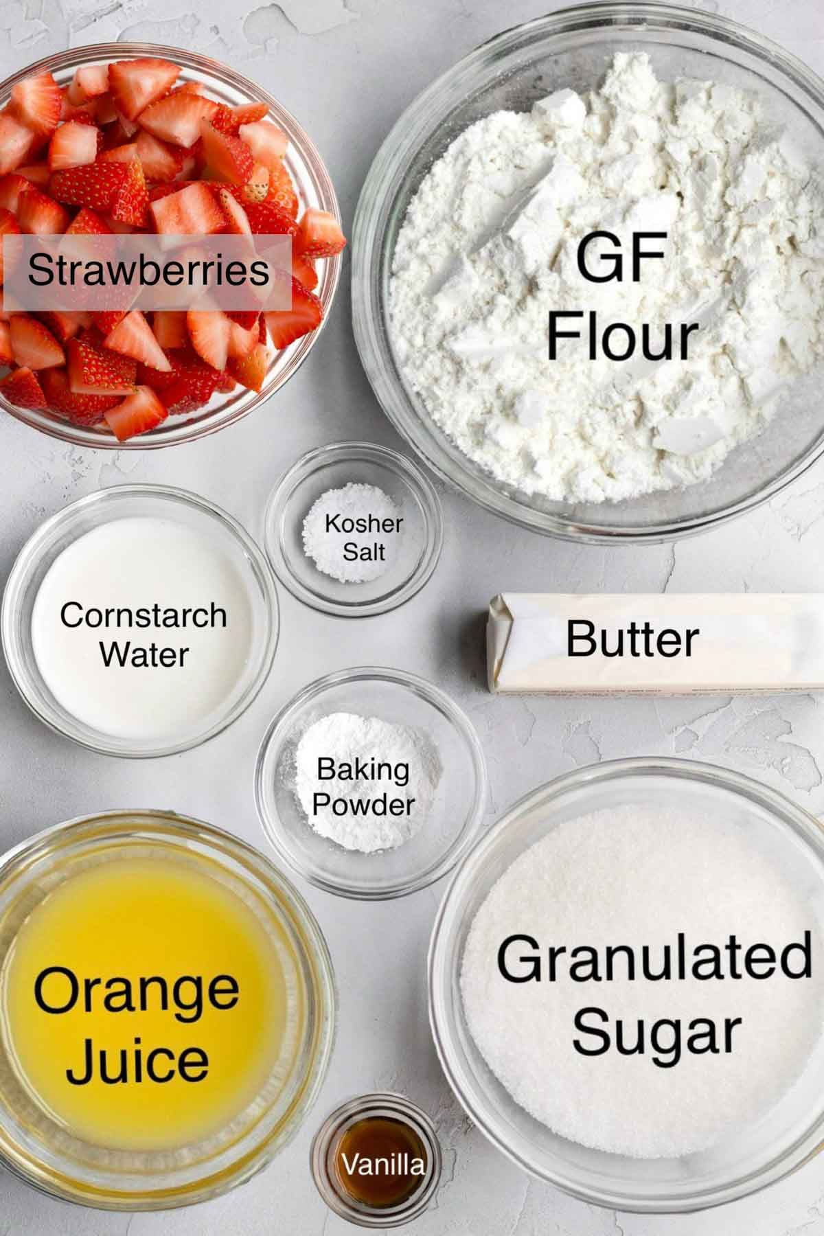 Strawberry cobbler ingredients in separate bowls: Strawberries, gluten free flour, cornstarch water. kosher salt, butter, baking powder, orange juice, granulated sugar, and vanilla.